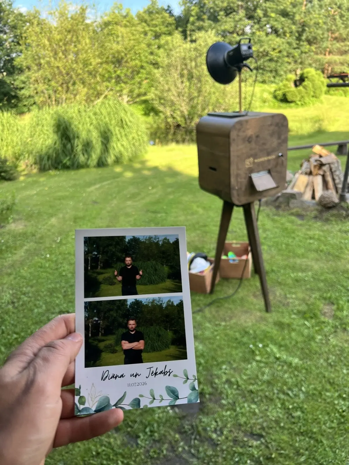 Rokā kāzu izdruka ar jaunā pāra vārdiem, fonā foto kaste dārzā