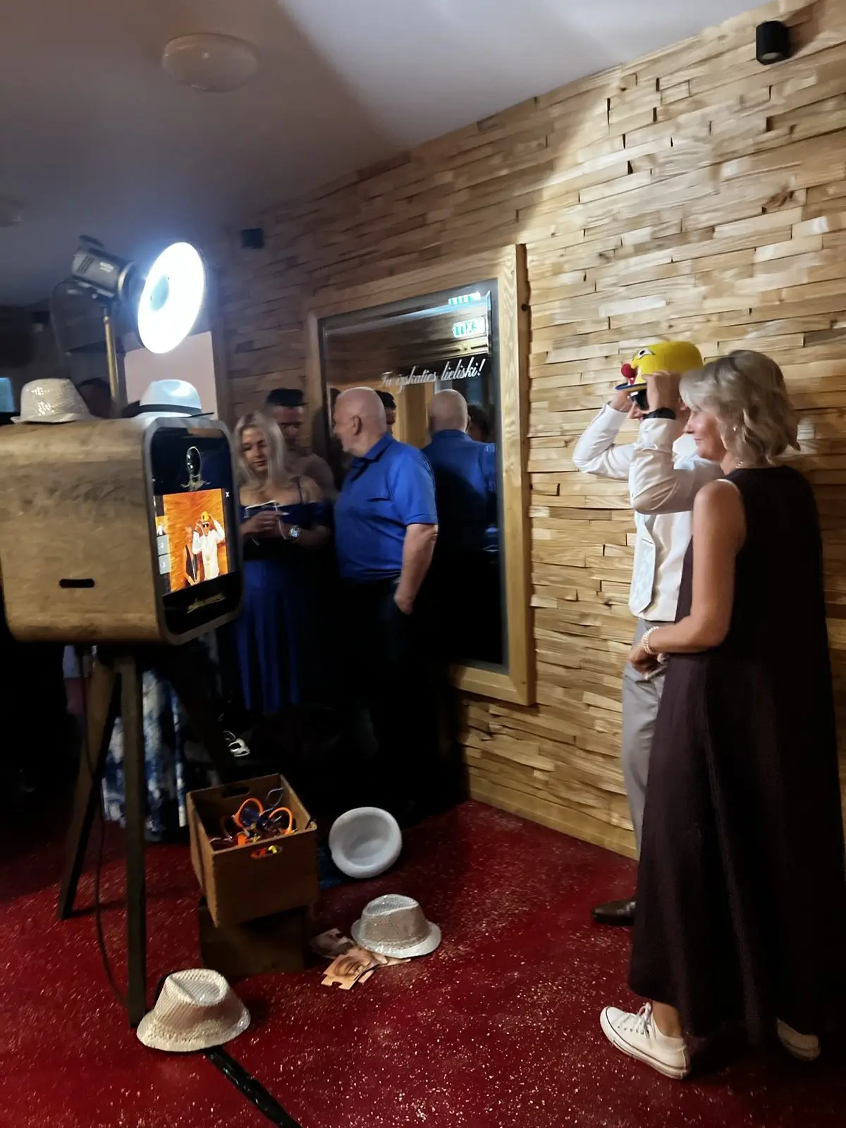 Viesi ar cepurēm un rekvizītiem stāv pie foto kastes ballītē